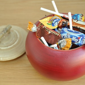 Puede incluir: Un cuenco de madera rojo con forma de manzana lleno de caramelos y piruletas envueltos. El cuenco tiene una tapa de madera con un asa de bambú.