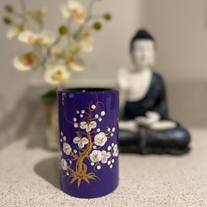 May include: A purple ceramic vase with a gold and white floral design. The vase is decorated with a stylized tree with white flowers and gold branches.