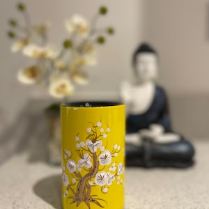 May include: A yellow ceramic vase with a white floral design. The vase has a black rim and a glossy finish.