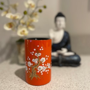 Könnte beinhalten: Orangefarbene Keramikvase mit schwarzem Rand und einem goldenen und weißen Blumendesign. Die Vase zeigt einen stilisierten Baum mit weißen Blüten und goldenen Ästen.