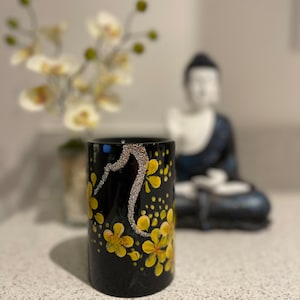 May include: Black cylindrical vase with a floral design featuring yellow flowers and gold accents. The vase is decorated with small gold dots and a silver swirl.