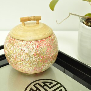 May include: A round, decorative jar with a mosaic design in shades of pink, white, and gold. The jar has a bamboo lid with a handle. The jar is sitting on a black tray with a silver reflective surface.