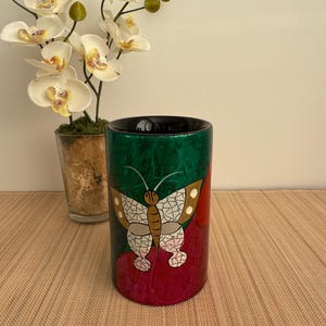 May include: A cylindrical ceramic vase with a green, red, and white butterfly design. The vase has a black rim and is decorated with a mosaic-like pattern.