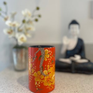 May include: A red cylindrical vase with a floral design. The vase is decorated with yellow flowers and a silver glitter-like design. The vase is sitting on a white countertop.