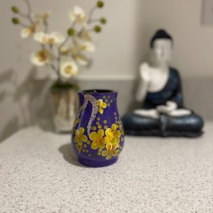 May include: A purple ceramic vase with a floral design featuring yellow flowers and white dots. The vase is sitting on a white countertop.