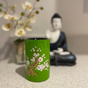 Könnte beinhalten: Ein grüner Keramikzylinder mit einem weißen und goldenen Blumendesign. Der Zylinder hat eine glänzende Oberfläche und ist mit einem stilisierten Baum mit weißen Blüten verziert.