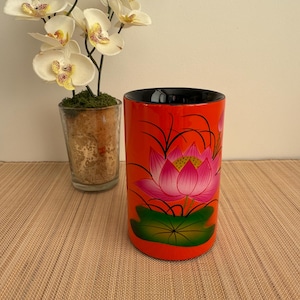 May include: A red cylindrical vase with a black rim, hand-painted with a pink lotus flower and green leaves. The vase is sitting on a brown woven mat.