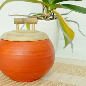 May include: Orange ceramic jar with a wooden lid and handle. The jar has a wood grain pattern and is sitting on a woven mat.