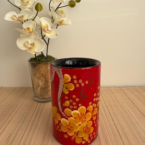 May include: A red cylindrical vase with a black rim and a floral design. The vase is decorated with yellow flowers and red dots on a red background.