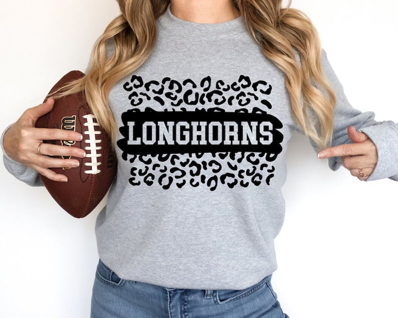 May include: A gray sweatshirt with a black leopard print design and the word "Longhorns" in white letters. The sweatshirt is being held by a person holding a football.