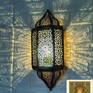 Puede incluir: Linterna ornamental de metal negro con intrincados patrones calados, que proyecta sombras decorativas sobre una pared de ladrillos blancos. La linterna tiene un diseño marroquí tradicional, con una luz cálida que emana del interior. La imagen incluye el logotipo "GatesofMorocco".