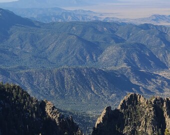 May include: A panoramic view of a mountain range with a blue sky and distant horizon. The mountains are covered in trees and have a rugged, rocky appearance. The text "the_nm_tx" is visible in the upper left corner of the image.