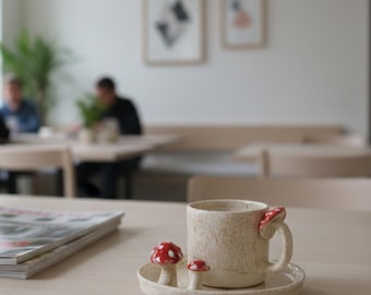 May include: A speckled ceramic mug with a mushroom-shaped handle and two mushroom figurines on a matching saucer. The mushrooms are red with white spots. The mug and saucer are beige.
