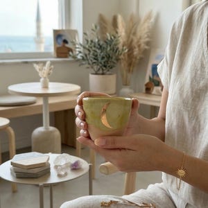 Op de afbeelding: Een handgemaakte keramische beker met een gouden halve maan ontwerp. De beker heeft een lichtgroene bovenkant en een perzikkleurige basis. De beker wordt in de handen van een persoon gehouden. Een gouden zon- en sterarmband is zichtbaar.