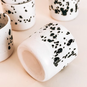 May include: A set of white ceramic mugs with black splatter paint. The mugs are stacked on top of each other and have a speckled design.