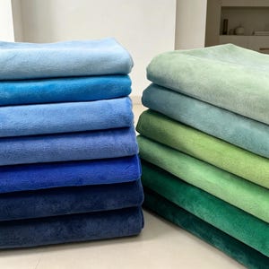 May include: Two stacks of folded plush fabric pieces arranged in a gradient color scheme. The left stack features shades of blue ranging from light sky blue to deep navy. The right stack displays shades of green from pale mint to dark forest green.