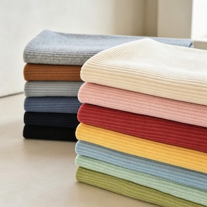 May include: A stack of folded ribbed knit blankets in various colors including cream, light pink, deep red, yellow, light blue, sage green, olive green, grey, brown, slate blue, and black, arranged in two neat columns on a flat surface.