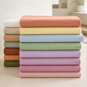 Könnte beinhalten: Ein Stapel gefalteter Stoffstücke in verschiedenen Unifarben, darunter Weiß, Beige, Pfirsich, Gelb, Hellblau, Salbeigrün, Lavendel, Mauve und Altrosa. Die Textilien sind ordentlich geschichtet und weisen eine glatte, matte Textur sowie einheitliche rechteckige Formen auf.