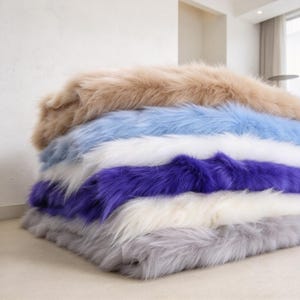 May include: A stack of six folded faux fur blankets in various colors, including tan, light blue, white, deep purple, cream, and light gray. Each blanket features a long, soft, shaggy texture layered neatly on top of one another in a vertical pile.