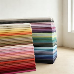 May include: A large stack of folded fabric pieces in a wide variety of colours, including shades of red, orange, yellow, green, blue, purple, pink, grey, and white. The textiles are neatly layered in two distinct columns, showing textures.