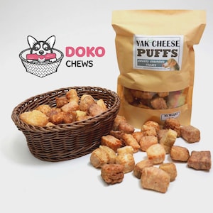 May include: A brown woven basket and a kraft paper bag filled with dog treats. The bag has a clear window and a label that reads "Yak Cheese Puffs". The treats are scattered around the basket and bag. The logo "DOKO CHEWS" is in the upper left.