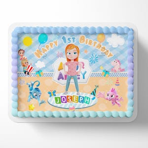 May include: A rectangular birthday cake with blue frosting and a decorative border. The cake features a cartoon illustration with the text "Happy 1st Birthday" and the name "Joseph". The design includes cartoon characters, balloons, and a sun.