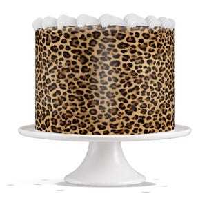 May include: A round cake with a leopard print design on a white cake stand. The cake is decorated with white frosting along the top edge. The cake stand has a pedestal base.