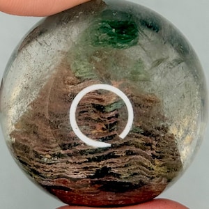 May include: A clear, round crystal ball with internal inclusions of brown, green, and white. The crystal has a layered appearance, with a white circular design on the surface. The ball is held between two fingers.