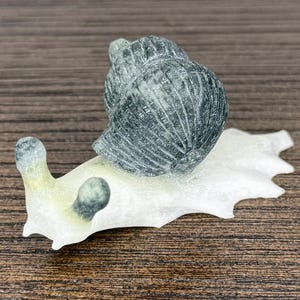 May include: A small snail figurine featuring a dark grey, textured, spiraled shell mounted on a translucent, pale white body. The snail has two prominent eye stalks with dark grey tips, showcasing a smooth, polished finish throughout the piece.