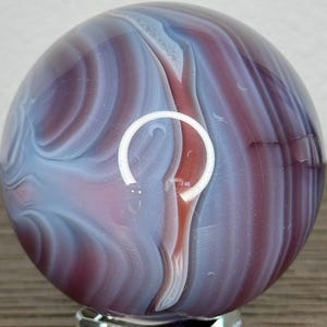 May include: A decorative glass marble with swirling patterns of blue, purple, and red. The marble is displayed on a clear, square glass stand. The colours blend together, creating a unique and artistic appearance.
