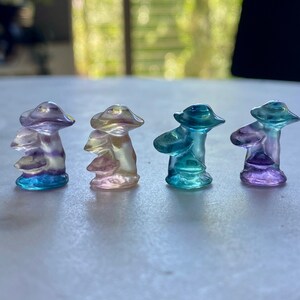Op de afbeelding: Vier kleine glazen paddenstoelfiguren in verschillende kleuren. De paddenstoelen zijn iriserend en hebben een gelaagde, gestapelde uitstraling. De kleuren zijn paars, roze, blauw en groen.