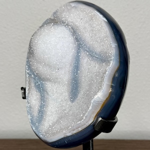 May include: A polished agate slice with a sparkling white druzy quartz center and dark blue outer banding, mounted on a black metal stand with a square base. The stone features a natural crystalline texture and smooth, dark edges.