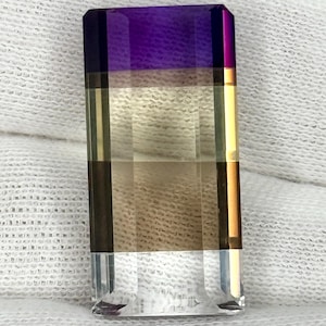 May include: A rectangular faceted gemstone featuring a color gradient that transitions from a deep purple at the top to a golden yellow in the center, fading into a light brown and ending in a clear, transparent base with sharp, clean edges.