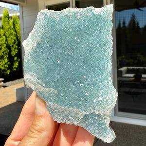 Può includere: Un grande gruppo di cristalli blu chiaro di forma irregolare con una superficie strutturata. Il cristallo ha un bordo smerigliato e cristallino. Il cristallo è tenuto nella mano di una persona, con uno sfondo sfocato di vegetazione e una casa.