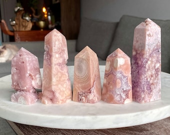 May include: Six pink and purple crystal points arranged on a white marble surface. The crystals are all different shapes and sizes, and they have a variety of patterns and textures.