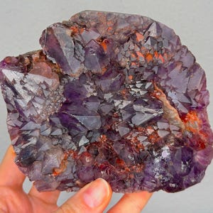 May include: A raw amethyst geode with a deep purple hue, featuring crystalline structures and reddish-orange accents. The natural formation is held in a hand, showcasing its irregular shape and texture. This unique specimen is a beautiful example of natural mineral art.