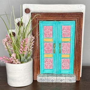 Könnte beinhalten: Eine dekorative Miniaturtür mit türkisfarbenen Doppeltüren, goldenen Akzenten und rosa gemusterten Paneelen. Die Tür ist in dunklem Braunholz gerahmt und in einer weißen, stuckartigen Struktur platziert. Links befindet sich ein kleiner weißer Pflanztopf mit rosa und grünen Kunstpflanzen.