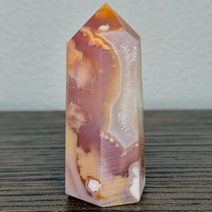 May include: A polished agate crystal point in shades of orange, pink, and white. The gemstone has a hexagonal shape and a pointed top. The crystal is displayed on a wooden surface.