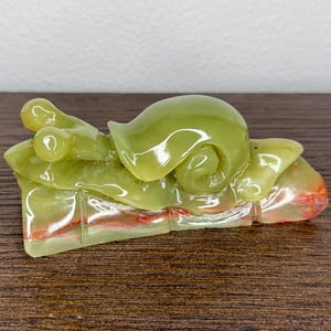 May include: A carved stone snail figurine in shades of light green and translucent olive, featuring a smooth, polished surface with a distinct spiral shell and two eye stalks, resting on a base with subtle reddish-orange mineral inclusions.