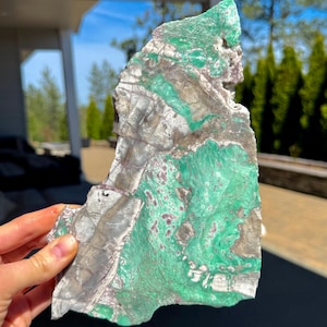 May include: A large, irregularly shaped slab of natural stone, featuring a striking blend of colors. The stone showcases a vibrant green hue, interspersed with areas of gray, brown, and hints of red. The surface has a textured appearance.