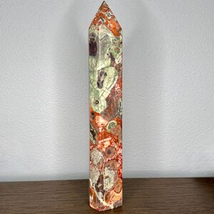 May include: A tall, polished crystal tower with a hexagonal shape. The stone features a complex, mottled pattern of deep red, burnt orange, and pale sage green colors, with visible natural inclusions and veins throughout the smooth surface.
