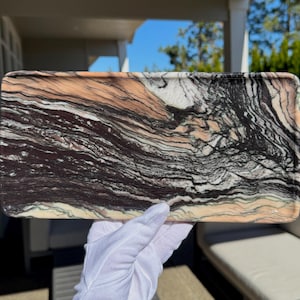 May include: A rectangular stone tray featuring a natural, swirling pattern of deep black, cream, and soft peach-coloured veins. The surface is smooth and polished, displaying intricate, flowing mineral layers that create a unique, organic aesthetic.