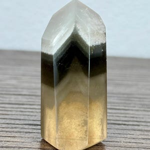 May include: A polished, multi-layered crystal tower featuring a pointed top. The stone displays distinct horizontal bands of color, transitioning from a clear, translucent apex to a dark charcoal-black middle section, ending in a warm, golden-yellow base.