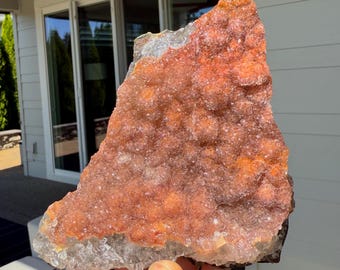Op de afbeelding: Een grote, ruwe kristalcluster met een kleurengradatie. De basis is een doorschijnend, lichtgrijs, dat overgaat in een levendige oranje en roodbruine tint. Het oppervlak is bedekt met kleine, sprankelende kristalformaties.