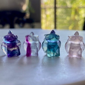 Puede incluir: Cuatro pequeñas figuras de búhos talladas en cristal de fluorita de colores. Los búhos son en tonos de morado, rosa, azul y verde. Cada búho tiene un cuerpo redondeado, alas y una cabeza puntiaguda. Las figuras se exhiben sobre una superficie clara.