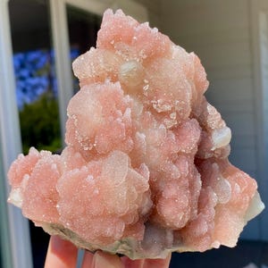 May include: A cluster of pink quartz crystals with a sugary texture. The crystals have a soft, pastel pink hue with areas of white and a slight sparkle. The quartz formation has a natural, organic shape, with multiple crystal points.