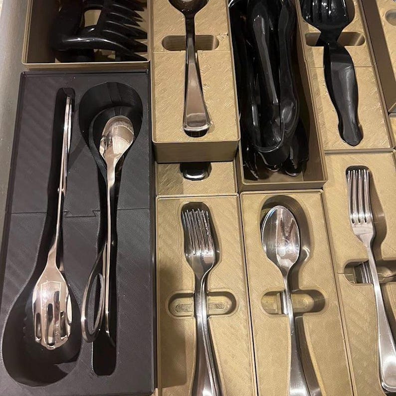 May include: A set of silverware, including spoons, forks, and serving utensils, neatly arranged in individual compartments. The silverware is a shiny silver color, and the compartments are a light brown color. The set appears to be new and unused.