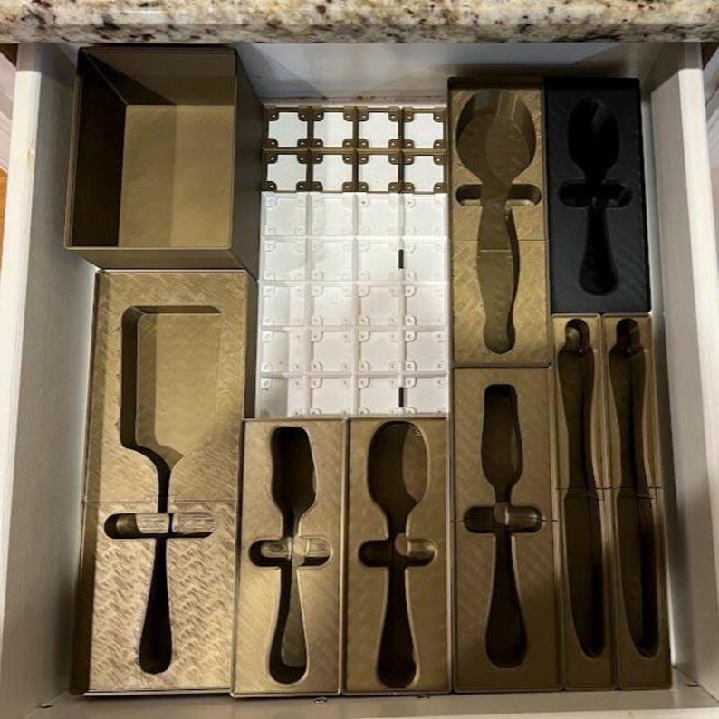 May include: A drawer organizer with compartments for kitchen utensils. The organizer includes gold-colored trays and black and white plastic dividers. The compartments are shaped to hold various utensils, such as spoons, knives, and spatulas.