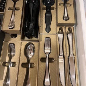 May include: A drawer filled with various silverware pieces, including forks, spoons, and knives. The silverware is silver-colored and neatly arranged in individual cardboard boxes. Some utensils are black.
