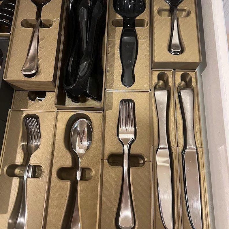 May include: A drawer filled with various silverware pieces, including forks, spoons, and knives. The silverware is silver-colored and neatly arranged in individual cardboard boxes. Some utensils are black.